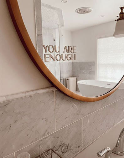 Modern bathroom with pink and grey marble tiles and a circular wooden mirror has an affirmation sticker that says you are enough
