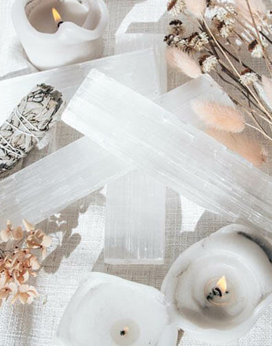 Scented lit candles burning next to clear quartz crystals, sage and dried flowers on dreamy table with white linen