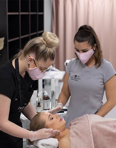 dermal therapist teaches student how to give client lying on treatment bed a facial using skin products and massage techniques
