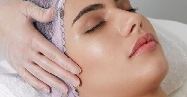 Dermal therapist gives client wearing a pink hair net a facial using a microdermabrasion pen to exfoliate dead skin