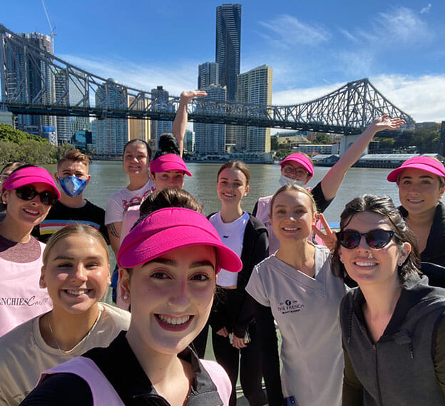group of students in pink hats participate in charity fun run along brisbane river and pose in front of bridge in the city