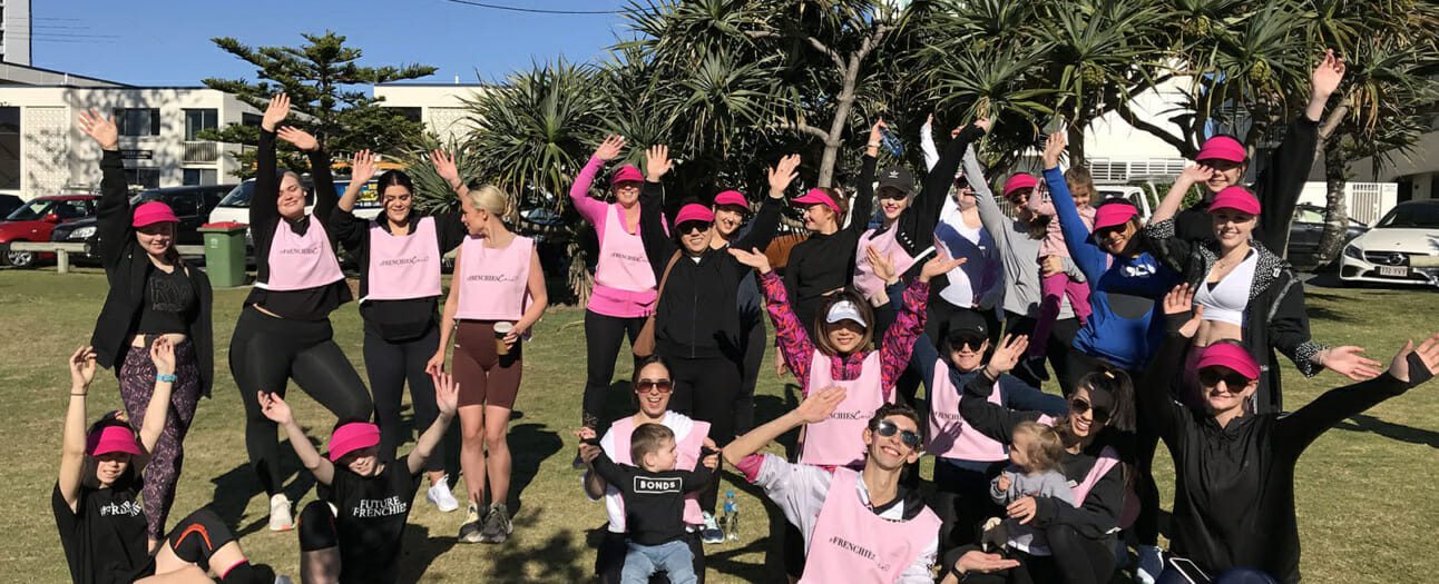 group of beauty students and families sit on grass in the sun dressed in pink activewear with their hands in the air after running for charity event