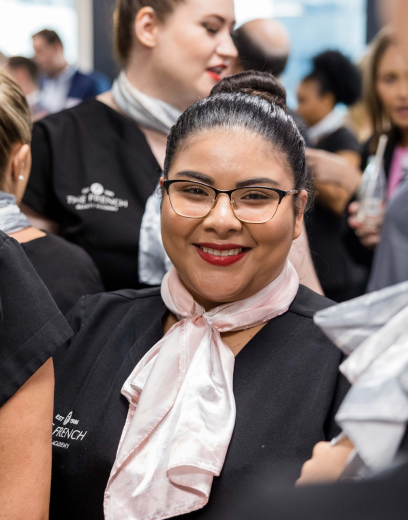 student with slicked back bun, red lipstick and black framed reading glasses is smiling at the camera wearing a pink scarf