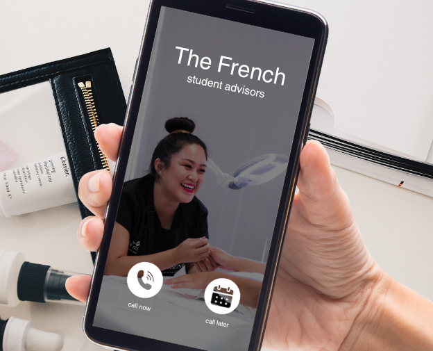 A hand holding an iPhone receiving a phone call from "The French Beauty Academy". The phone wallpaper shows a smiling woman giving a manicure treatment. Behind the phone is a picture of a desk with beauty products and a paperwork strewn about.