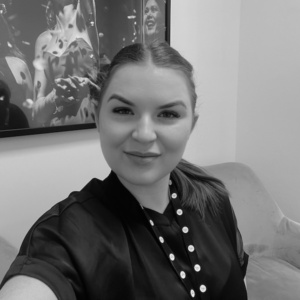 A black and white image of a woman dressed in a professional corporate top and wearing a lanyard taking a selfie in an office. The background shows an image of women celebrating as conteffi falls from the air hung on the wall.