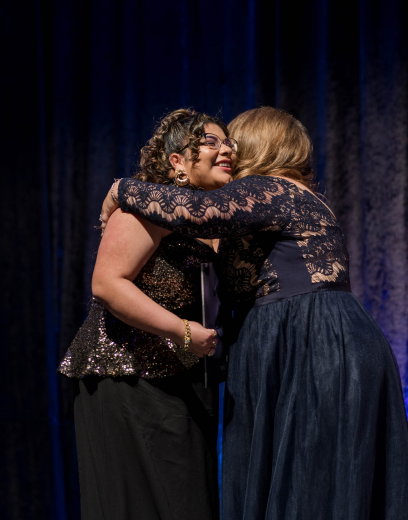 two women wearing formal gowns embracing on stage at a graduation ceremony event