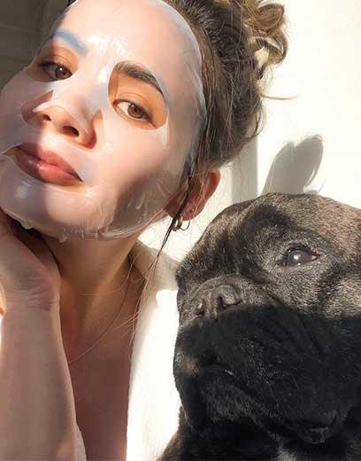 A girl wearing a sheet mask with her hair in a messy bun taking a selfie in golden hour sunlight with her pug dog