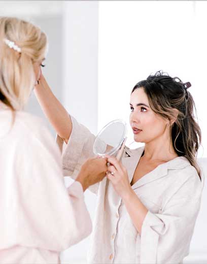 A blonde woman who's face you cannot see looks at herself in a handheld mirror as a brunette woman wearing her hair in a half up messy bun inspects her client's face in a skin care and eyebrow treatment consult