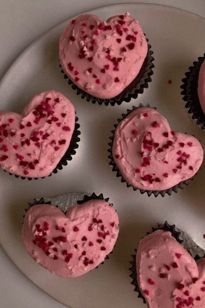 A plate of pink heart shaped cupcakes covered in red and pink heart sprinkles