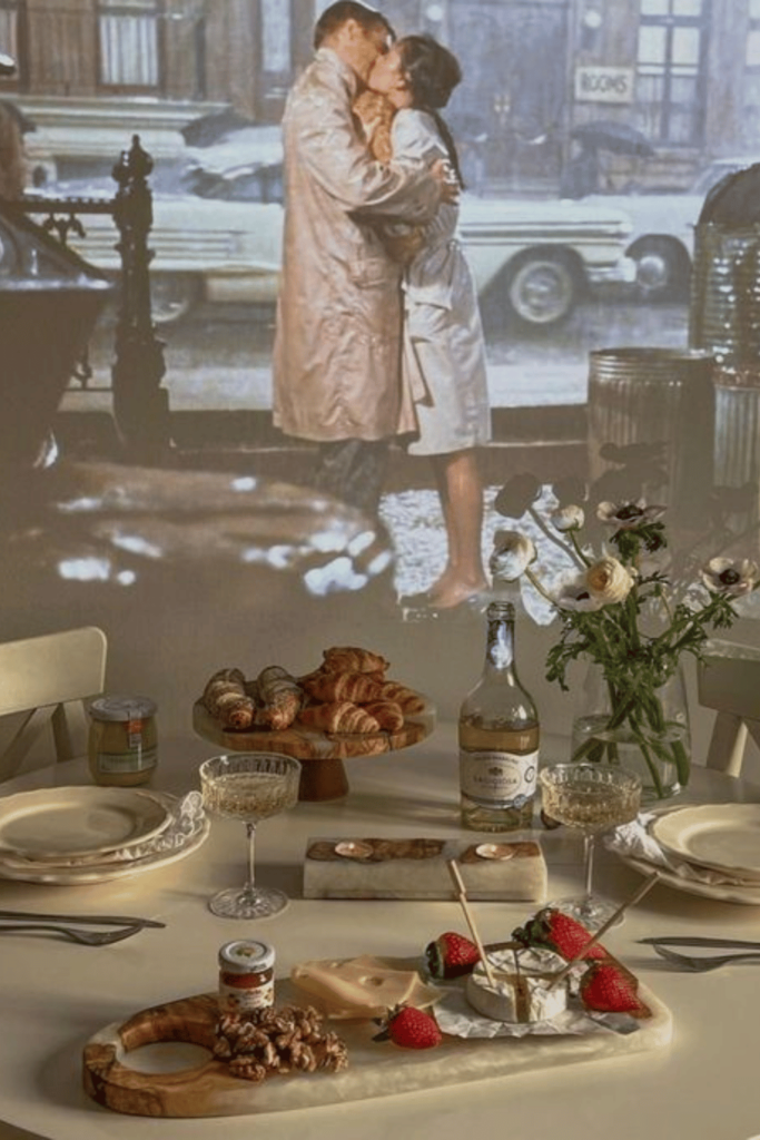 Breakfast At Tiffany's movie scene is projected onto a wall behind a romantic table setting. On the table is a cheese board, champagne, flowers, croissants and two place settings