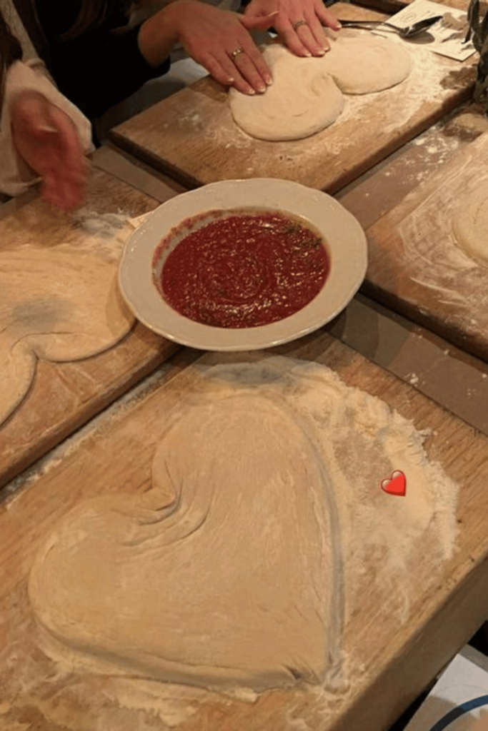 The scene depicts four friends making heart shaped pizza dough. In the centre of the table is a bowl of tomato paste