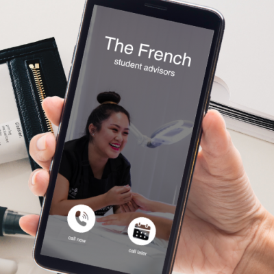 A hand holding an iPhone receiving a phone call from "The French Beauty Academy". The phone wallpaper shows a smiling woman giving a manicure treatment. Behind the phone is a picture of a desk with beauty products and a paperwork strewn about.